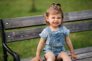 Adorable little girl laughing. Beautiful female kid in denim sitting on the bench in park. Happy little girl smile. Eighteen month