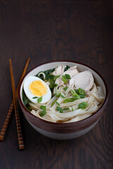 Chinese ramen soup made of chicken broth and noodles decorated with meat slices and halved egg in a bowl on dark brown wooden table together with chopsticks. Vertical orientation