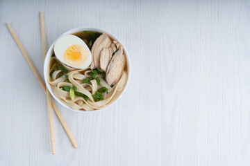 Asian spicy ramen soup made of chicken broth with noodles decorated by halved egg and meat slices in a bowl standing on white wooden table together with chopsticks. Image with copy space
