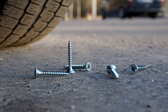 Several screws lie on the road, one stands upright in front of the car wheel. Wheel puncture concept.