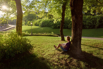 A woman sits on the grass under a tree on the edge of the park and reads a book at sunset.