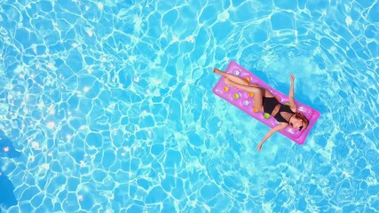 Fitted pretty girl in bikini chilling on inflatable pink mattress in swimming pool. Shaped woman in swimsuit tanning. Female relaxing on float in blue water at luxury resort. Aerial, view from above.