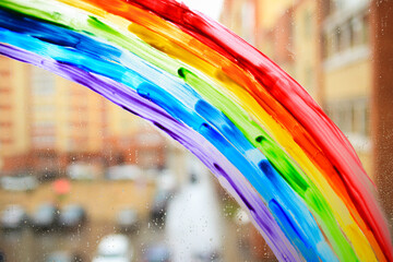 The rainbow is painted on the window. There are raindrops on the window and a city landscape. Bright painting on glass, gouache