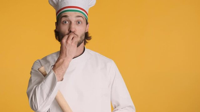 Attractive professional italian chef in uniform  emotionally showing bellissimo gesture and rolling pin on camera over colorful background