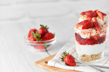 Homemade layered dessert with fresh strawberries, cream cheese or yogurt, granola and strawberry jam in glasses on white wood background. Healthy organic breakfast or snack concept. Selective focus.