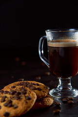 Close up of a glass cup of coffee, chocolate chip cookies and roasted coffee beans on dark background. Concept of ready to eat food, tasty snack. Selective focus, copy space.