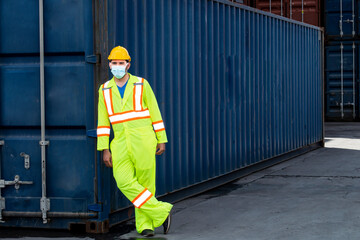 Smart engineer man wearing safety reflective cloth, helmet and surgical mask standing leaning on containers box from cargo freight ship for import and export at business logistics site in warehouse.