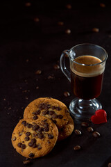 Close up of a glass cup of coffee, chocolate chip cookies and roasted coffee beans on dark background. Concept of ready to eat food, tasty snack. Selective focus, copy space.