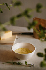 Pretty small ivory bowl with couscous porridge on table in the kitchen with cheese, spices and mint leaves decoration. Tasty and healthy vegetarian food isolated close up in rustic style