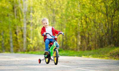 Boy kid training to ride bicycle with supporting wheels on sides, background summer park