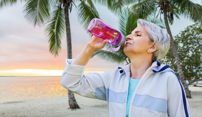 fitness, sport and healthy lifestyle concept - thirsty senior woman drinking water from bottle after exercising over tropical beach background in french polynesia