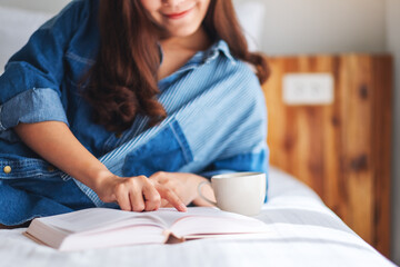 A beautiful young woman reading book and drinking hot coffee in a white cozy bed at home