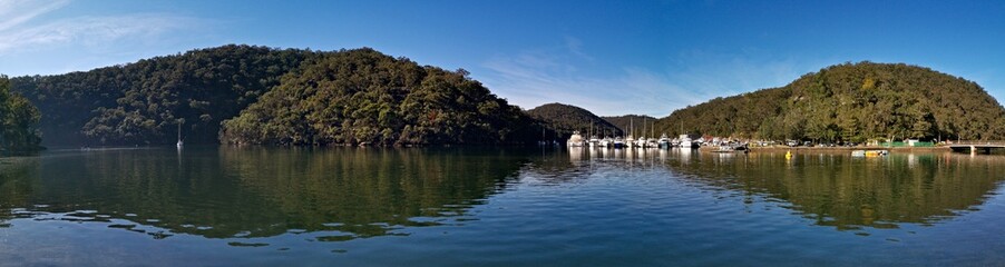 Fototapeta na wymiar Early morning view of a creek with beautiful reflections of blue sky, luxury boats, mountains and trees on water, Cowan Creek, Bobbin Head, Ku-ring-gai Chase National Park, New South Wales Australia