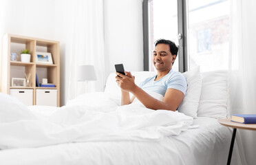 technology, bedtime and rest concept - happy smiling indian man with smartphone lying in bed at home