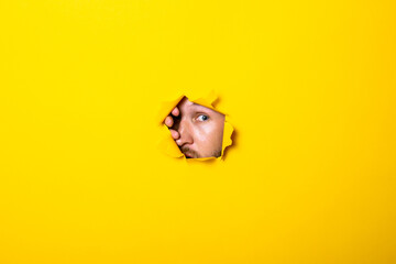 Young man looks with an eye into a torn hole in a yellow background