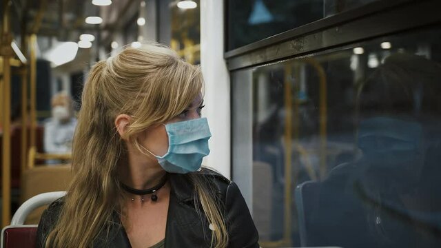 Female hipster in protective mask is sitting in moving tram and looking out the window. Covid-19 pandemic. Slow motion, dramatic cinematic portrait