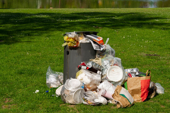 mit Abfall und Plastik Verpackungen &uuml;bervoller M&uuml;lleimer steht auf gr&uuml;ner Wiese. Symbolbild f&uuml;r Umweltverschmutzung.