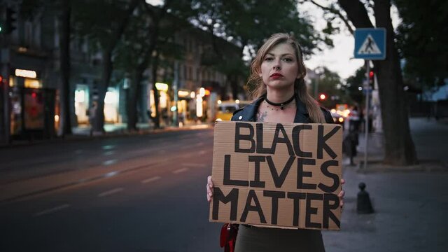 Girl hipster is holding cardboard tablet, inscription black lives matter. Standing by road against street with busy evening traffic. Dark cinematic