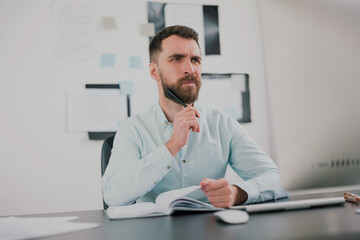 young bearded brunette man looking thoughtful while working on business project in his modern office, holding pen in his hand, work routine concept