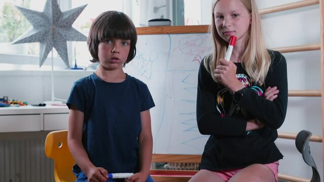 Brother and sister looking at the camera while sitting near the flipchart in home education. Gaining knowledge in self-isolation. Independent children.