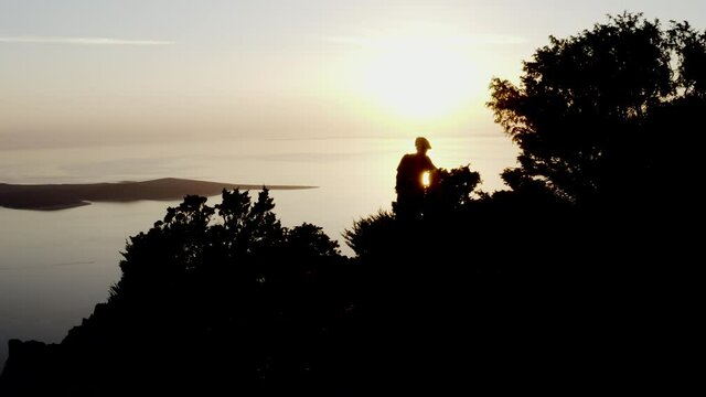 Gimbal shot of a silhouette of a fit mtb mountain bike rider, happy man on top of island walking with his bike after ride to top, sun shines flares into the camera trough threes Losinj Island, Croatia