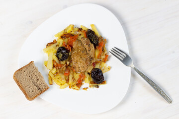 Meat stew with prunes garnished with fried potapo in plate on white wooden background with bread