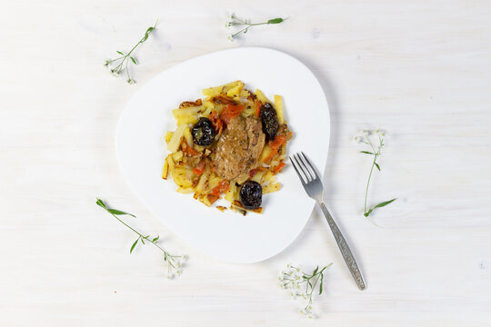 Meat stew with prunes garnished with fried potapo in plate on white wooden background decorated with flowers