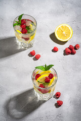 Fresh cold ice water drink with lemon, raspberry fruits and mint leaf in two faceted glass on stone concrete background, summer diet beverage, angle view