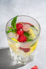 Fresh cold ice water drink with lemon, raspberry fruits and mint leaf in faceted glass on stone concrete background, summer diet beverage, angle view macro