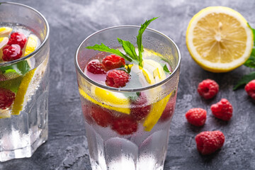 Fresh cold ice water drink with lemon, raspberry fruits and mint leaf in two faceted glass on stone concrete background, summer diet beverage, angle view selective focus
