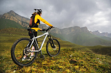 a girl in a helmet and with a backpack with a bicycle stands against the backdrop of epic mountains and rocks and cloudy evening sky. Cyclist looks at mountains before mtb descent