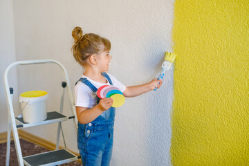 Cute little toddler girl painting the wall with yellow color in new house. Family repair apartment home. Happy child girl paints the wall, choosing color with palette, having fun with brush, indoors
