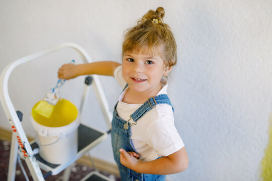 Adorable little toddler girl painting the wall with color in new house. Family repair apartment home. Happy baby child paints the wall, choosing color with palette, having fun with brush, indoors