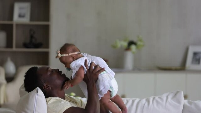 loving affectionate african dad playing and kissing infant mixed race daughter. Spbi father and little kid lifting and holding child. family bonding with toddler. lying on sofa indoors. concept happy