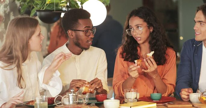 Crop view of multiethnic people smiling and chatting while spending time together. Millennial friends eating food and having conversation while sitting at table in cosy restaurant.