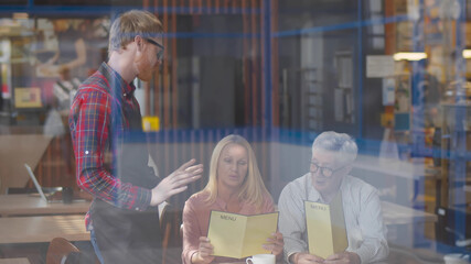 View through window of waiter helping senior couple with menu to choose dish in cafe