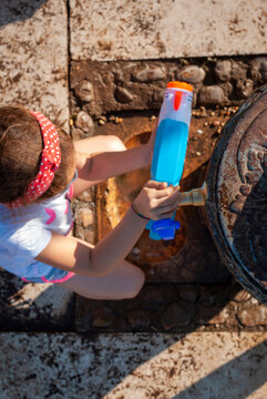 Summer in the city: little girl loads the water pistol at a fountain in a park