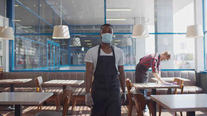 Fototapeta na wymiar Portrait of african waiter in apron safety mask and glves walking in cafe