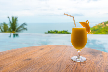 mango smoothie with a slice of mango stands on the table against the background of the pool, open air restaurant, glass with juice