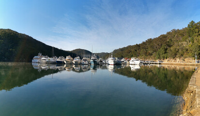 Fototapeta na wymiar Early morning view of a creek with beautiful reflections of blue sky, luxury boats, mountains and trees on water, Cowan Creek, Bobbin Head, Ku-ring-gai Chase National Park, New South Wales Australia