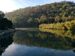 Fototapeta premium Early morning view of a calm creek with beautiful reflections of blue sky, mountains and trees on water, Cockle Creek, Bobbin Head, Ku-ring-gai Chase National Park, New South Wales Australia