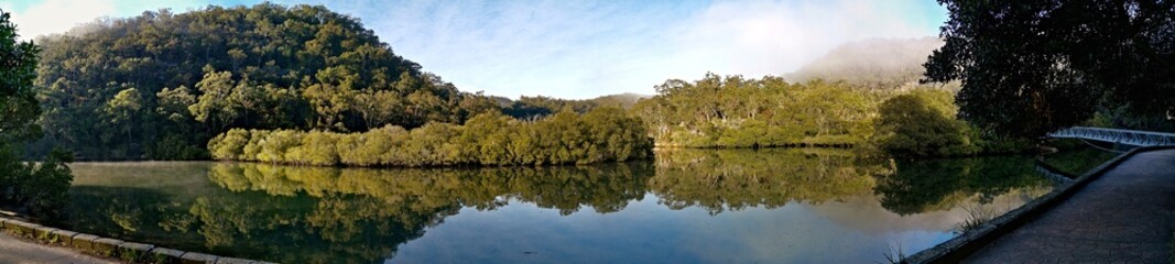 Fototapeta na wymiar Early morning panoramic view of a creek with beautiful reflections of blue sky, fog, mountains and trees on water, Cockle Creek, Bobbin Head, Ku-ring-gai Chase National Park, New South Wales Australia