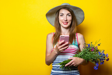 Smiling woman in hat with flowers holding phone on yellow background.
