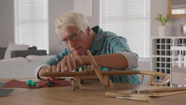 Focused senior man using sandpaper to polish wooden airplane sculpture. Mature carpenter working on making wooden flight at home. Happy old grandfather at home busy making handmade sculpture.