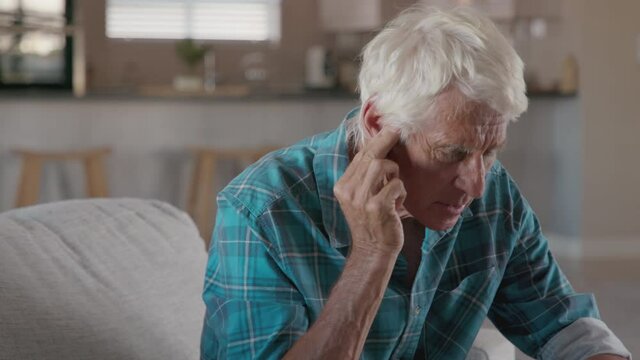 Senior man holding ears while suffering from pain. Tensed mature man sitting at home holding ears. Side view of senior man with symptom of hearing loss, copy space. Mature retired man in pain.