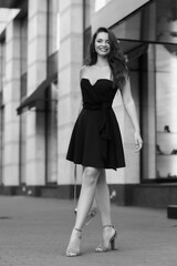 Young beautiful stylish woman in vlack dress, shoes, sunglasses holding handbag standing and posing at city street. Full length outdoor portrait