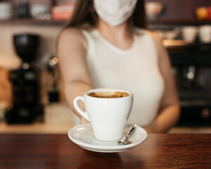 Barista girl in protective mask gives a cup of coffee to a cafe client