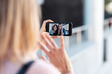 Back view of woman having video conversation with two pretty young business women while staying in the street.