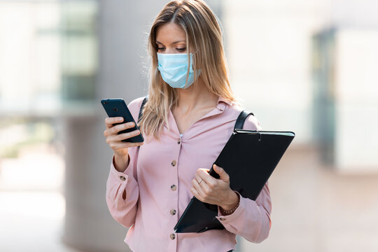 Beautiful Blonde Woman In Face Mask Using Her Mobile Phone While Walking In The Street.