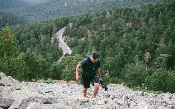 Senderista con bastones en primer plano subiendo por canchal hacia la Bola del Mundo con carretera de montaña al fondo en el Parque Nacional de la Sierra de Guadarrama en Madrid